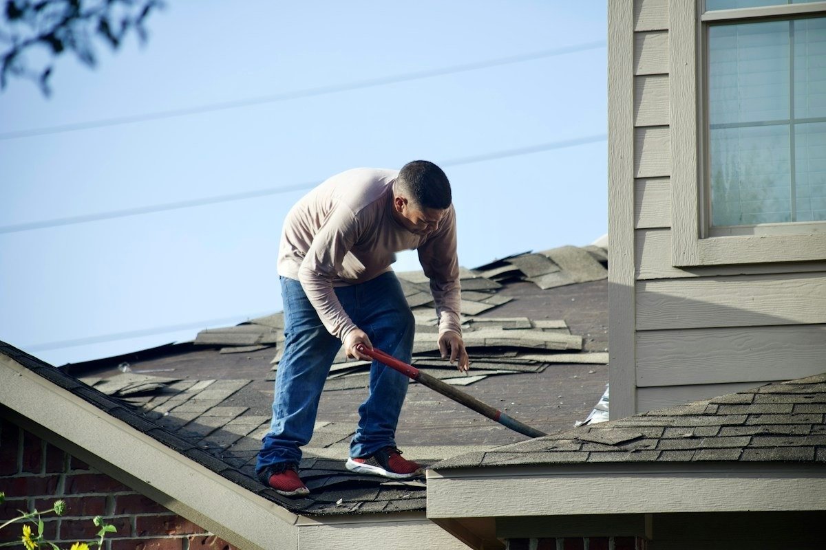 A roofer prying off worn asphalt shingles with a long-handled shovel