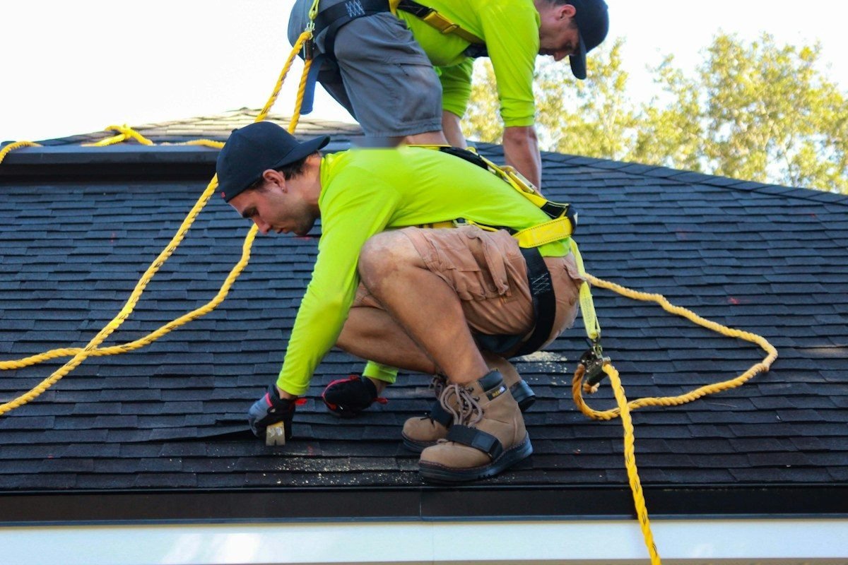 Two roofers in safety harnesses and bright shirts working on a dark shingle roof