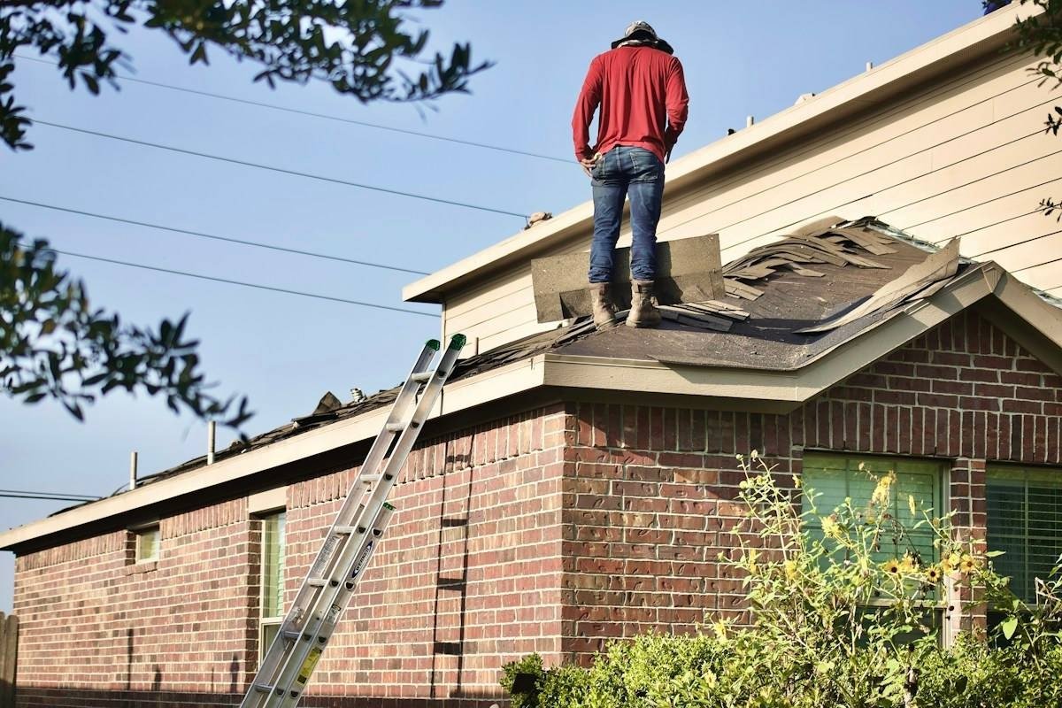 A roofer standing on a brick ranch house during a tear-off, old shingles piled on the roof and a ladder against the wall